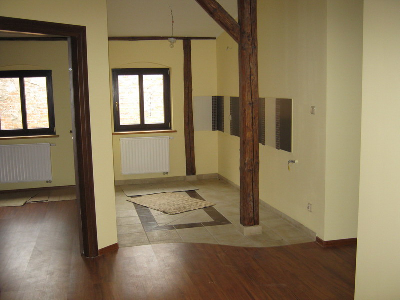 Wnętrze remontowanego pomieszczenia z drewnianą kolumną, fragmentem nowej podłogi laminowanej i położonymi płytkami podłogowymi, widoczne grzejniki i okna.